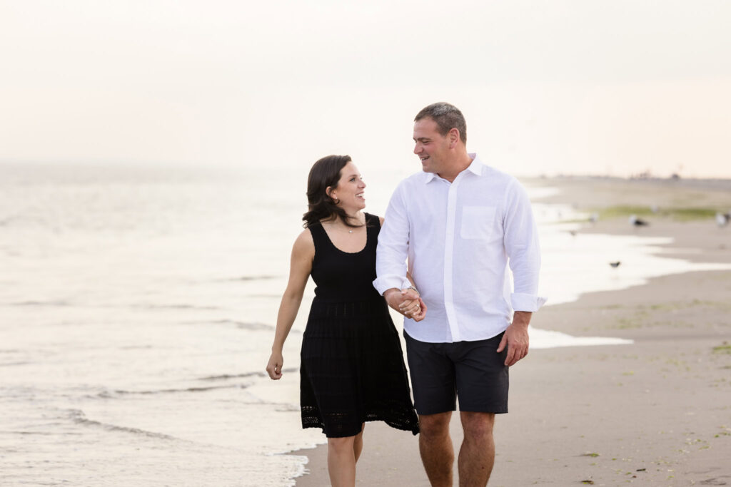 Nina and Scott holding hands and smiling as they walk along the shoreline at Breezy Point during their engagement session.