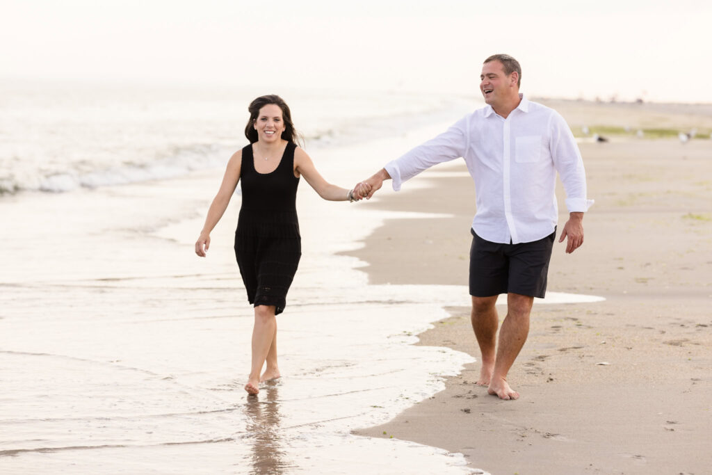 Engaged couple walking barefoot and laughing along the shoreline at Breezy Point Beach