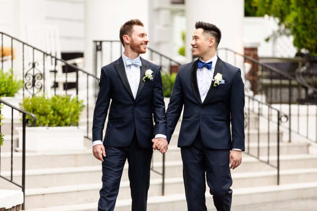 Two grooms walking hand-in-hand and smiling outside The Briarcliff Manor during their spring wedding day.