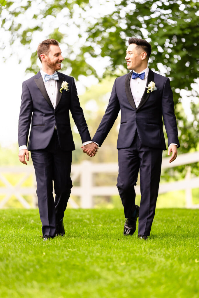 Two grooms holding hands and walking across the green lawn at The Briarcliff Manor during their spring wedding.