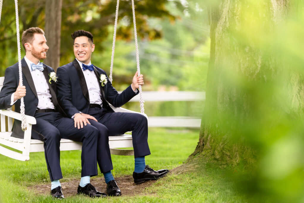Grooms sitting together on a white swing beneath a tree at The Briarcliff Manor, sharing a lighthearted moment.