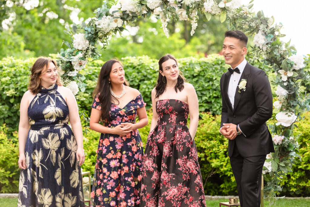 One groom standing at the floral arch with bridesmaids during the ceremony at The Briarcliff Manor, all smiling warmly.
