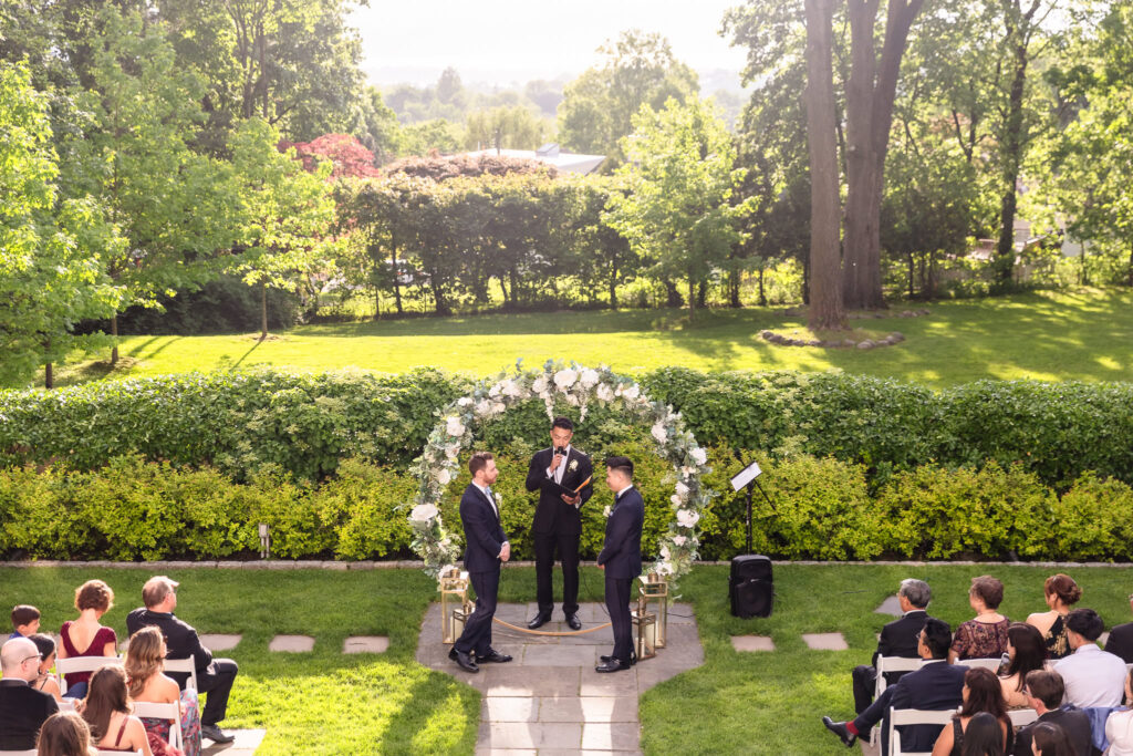 Outdoor spring wedding ceremony at The Briarcliff Manor, with two grooms standing beneath a floral arch as guests look on under golden afternoon light.