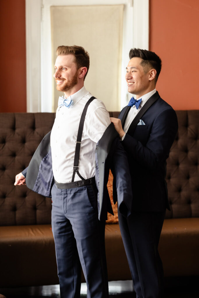 One groom helps the other into his jacket in the groom’s suite at The Briarcliff Manor, sharing a joyful moment before the ceremony.