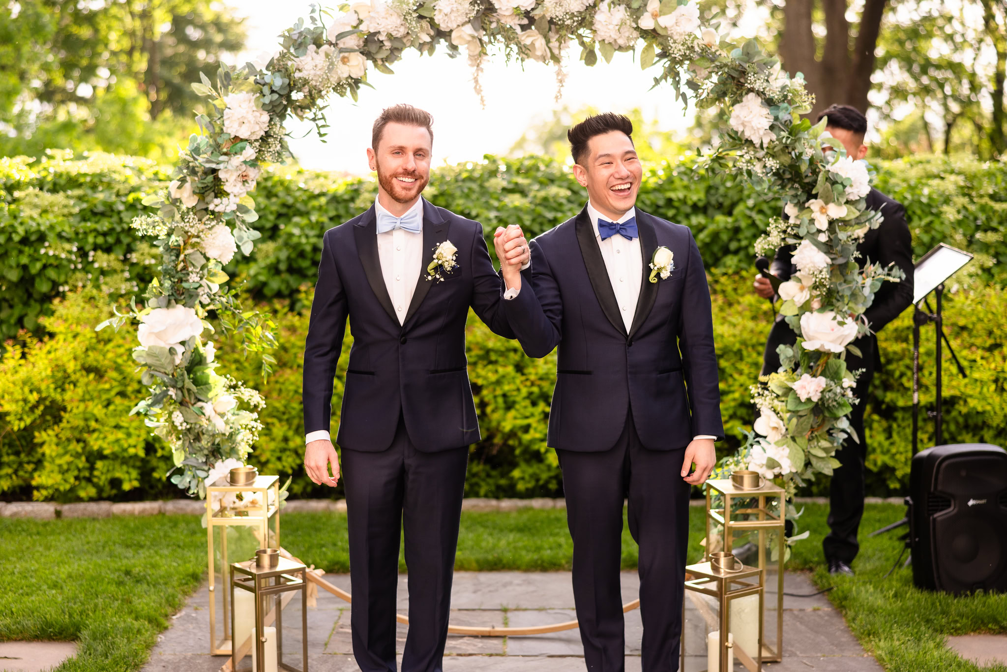 Groom and groom holding hands and smiling beneath a floral arch after their spring wedding ceremony at The Briarcliff Manor.