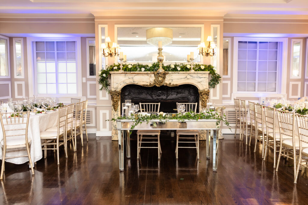 Sweetheart table set in front of the grand fireplace in The Briarcliff Manor ballroom, surrounded by candlelit reception tables.

