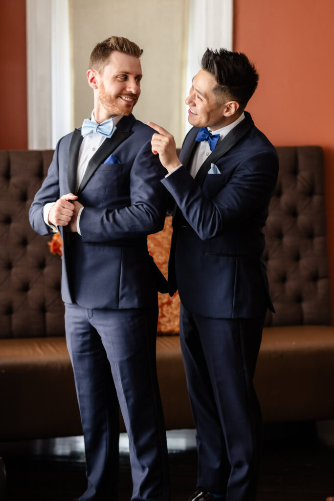 Groom adjusting his partner’s bow tie as they smile at each other in the groom’s suite at The Briarcliff Manor.