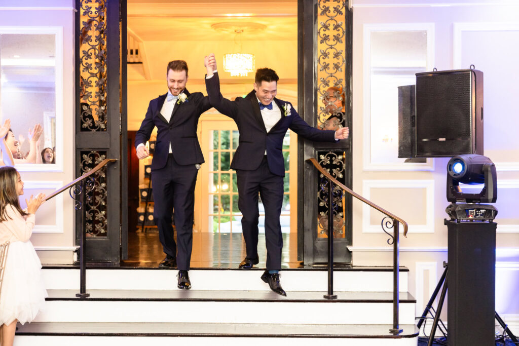 Grooms entering their wedding reception at The Briarcliff Manor hand-in-hand, smiling and cheered on by guests.
