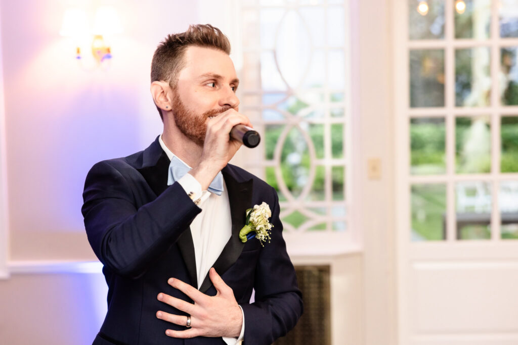 Groom holding a microphone and singing during the wedding reception at The Briarcliff Manor, hand over his heart.