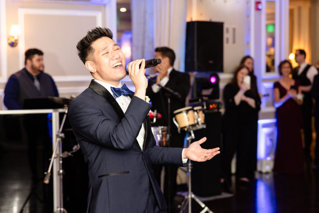 Groom singing passionately into a microphone during the reception at The Briarcliff Manor, with live musicians in the background.