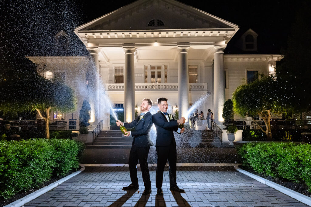 Grooms popping champagne and spraying it joyfully in front of The Briarcliff Manor’s illuminated facade at night.