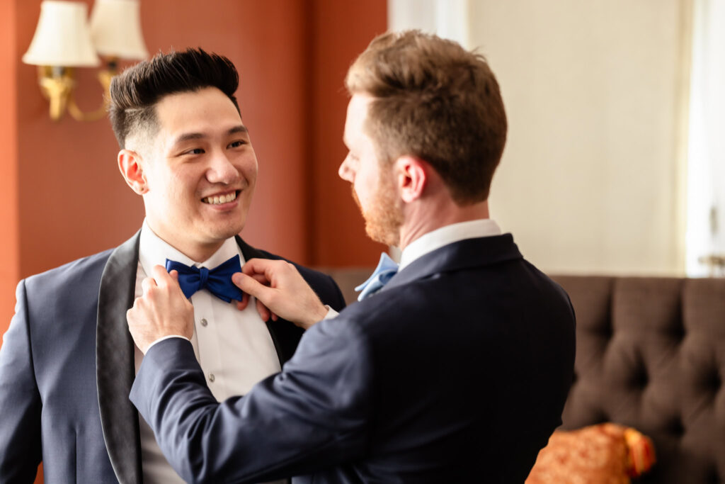 One groom adjusts his partner’s blue bow tie as they smile at each other during their wedding morning at The Briarcliff Manor.