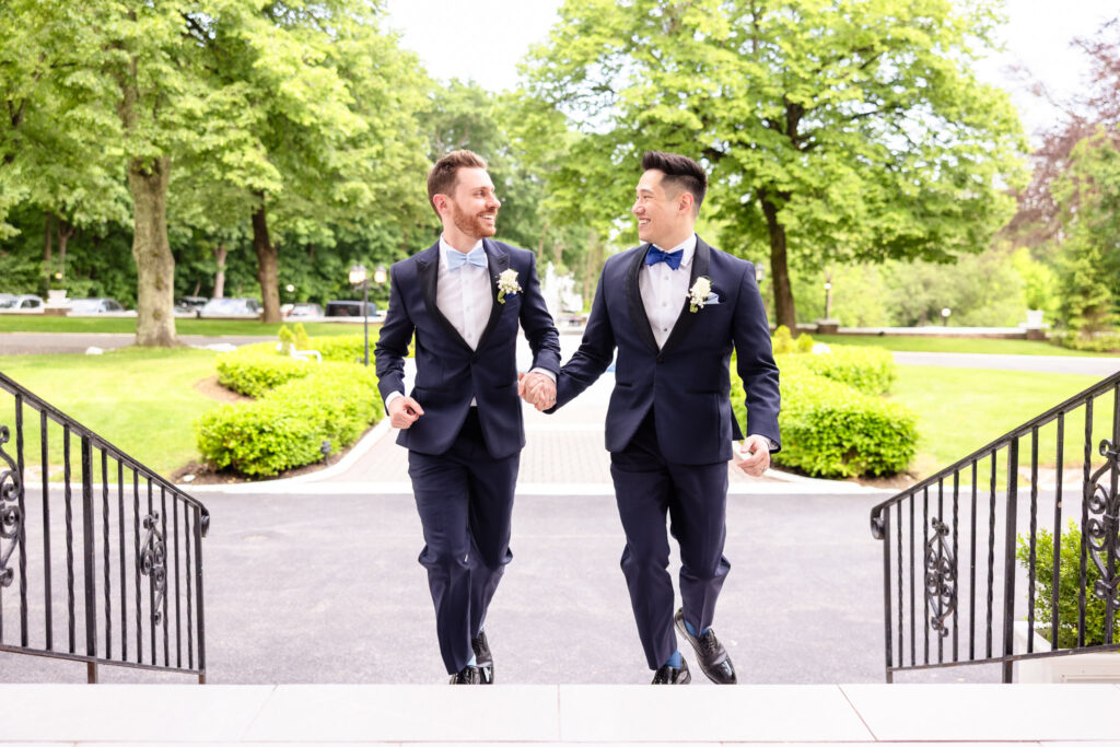 Two grooms walking hand-in-hand up the front steps of The Briarcliff Manor, smiling at each other on their wedding day.