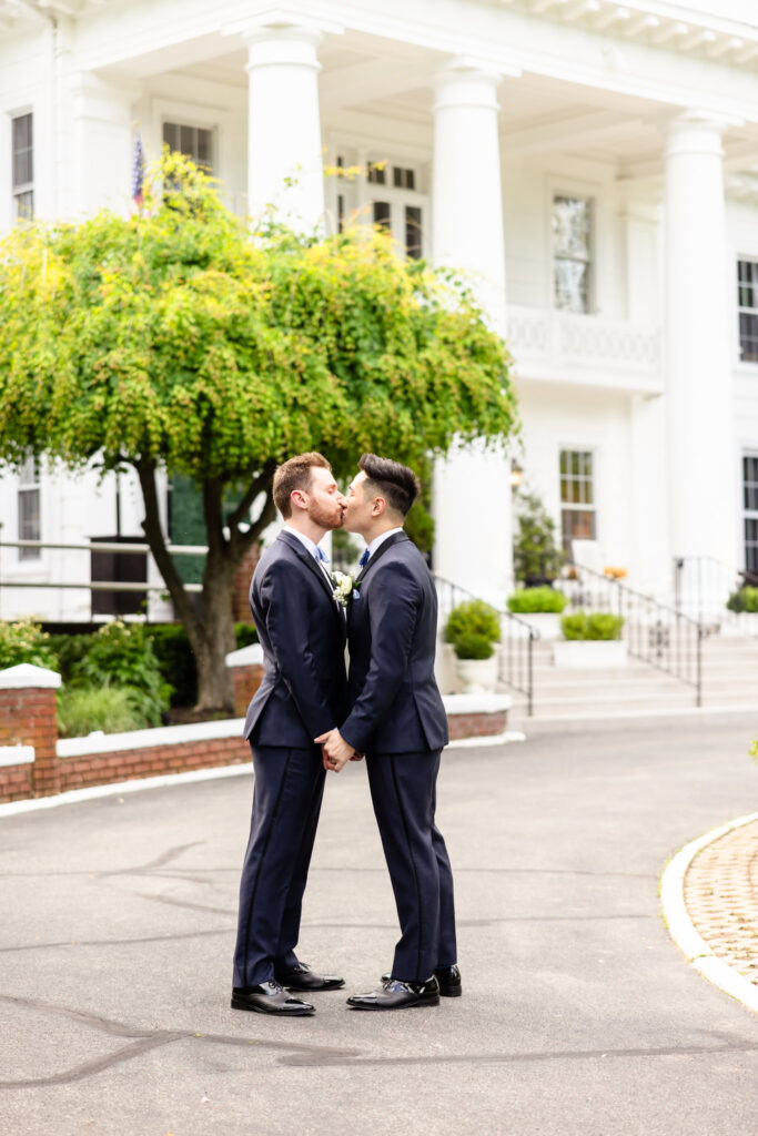 Grooms sharing a kiss in front of The Briarcliff Manor’s white-columned entrance beneath a spring-green tree.