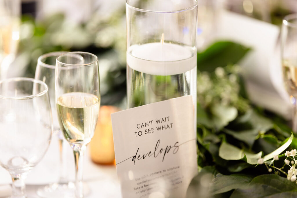 Close-up of a wedding reception table with champagne glasses, floating candle, greenery, and a sign reading “Can’t wait to see what develops.”