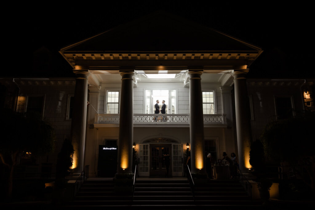 Silhouette of two grooms standing on the upper balcony of The Briarcliff Manor at night, framed by glowing white columns and architectural lighting.