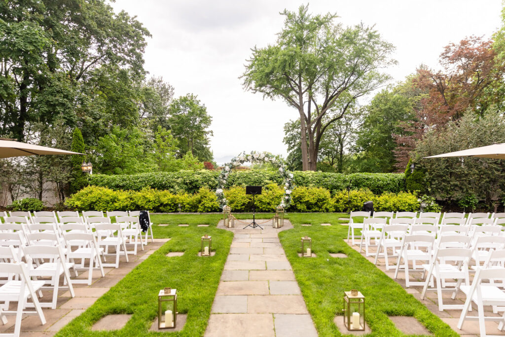 Outdoor garden ceremony setup at The Briarcliff Manor with white chairs, floral arch, and stone aisle lined with lanterns.