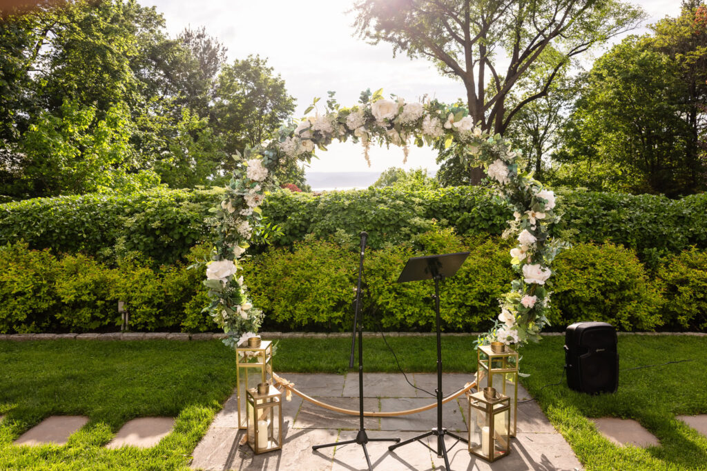 Sunlit floral wedding arch with white roses and greenery set against the garden hedges at The Briarcliff Manor.