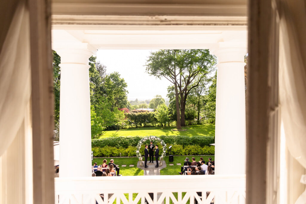 View from the balcony of The Briarcliff Manor overlooking an outdoor wedding ceremony in the garden with lush greenery and a floral arch.