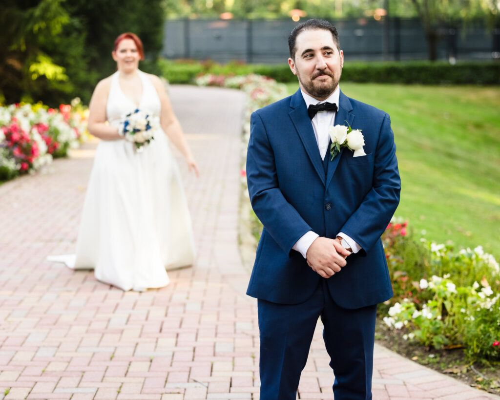 Groom stands with anticipation as the bride approaches for their first look along a flower-lined path at Crest Hollow Country Club.

