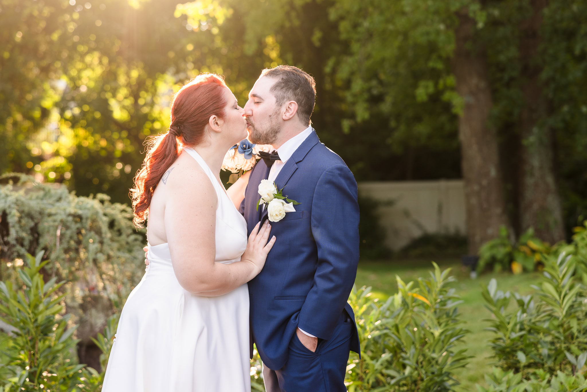 A bride and groom share a soft kiss in a sun-drenched garden at Crest Hollow Country Club, framed by lush greenery and golden light.