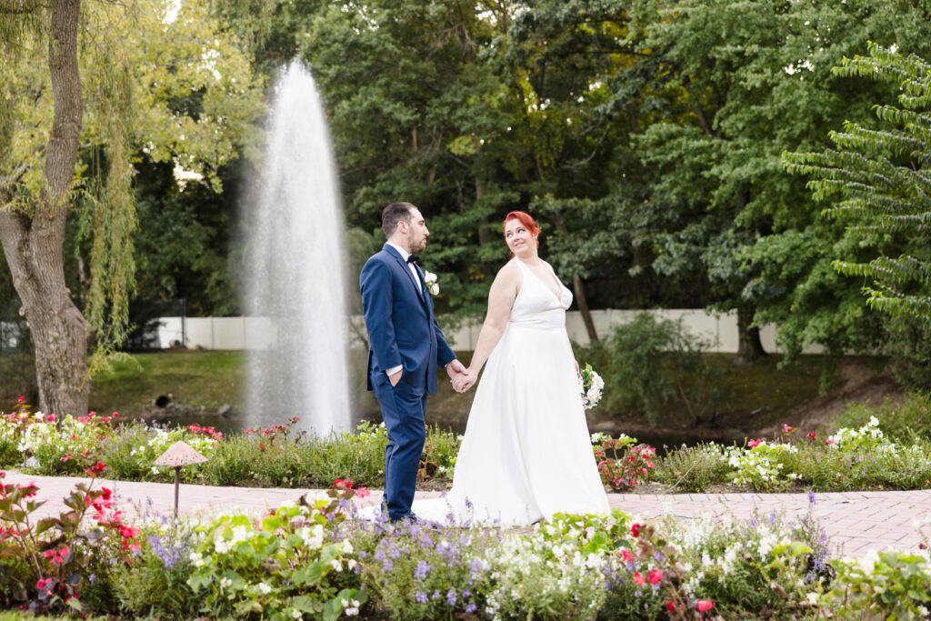 Bride and groom walk hand-in-hand through a vibrant garden at Crest Hollow Country Club, with a romantic fountain in the background and floral blooms surrounding them.