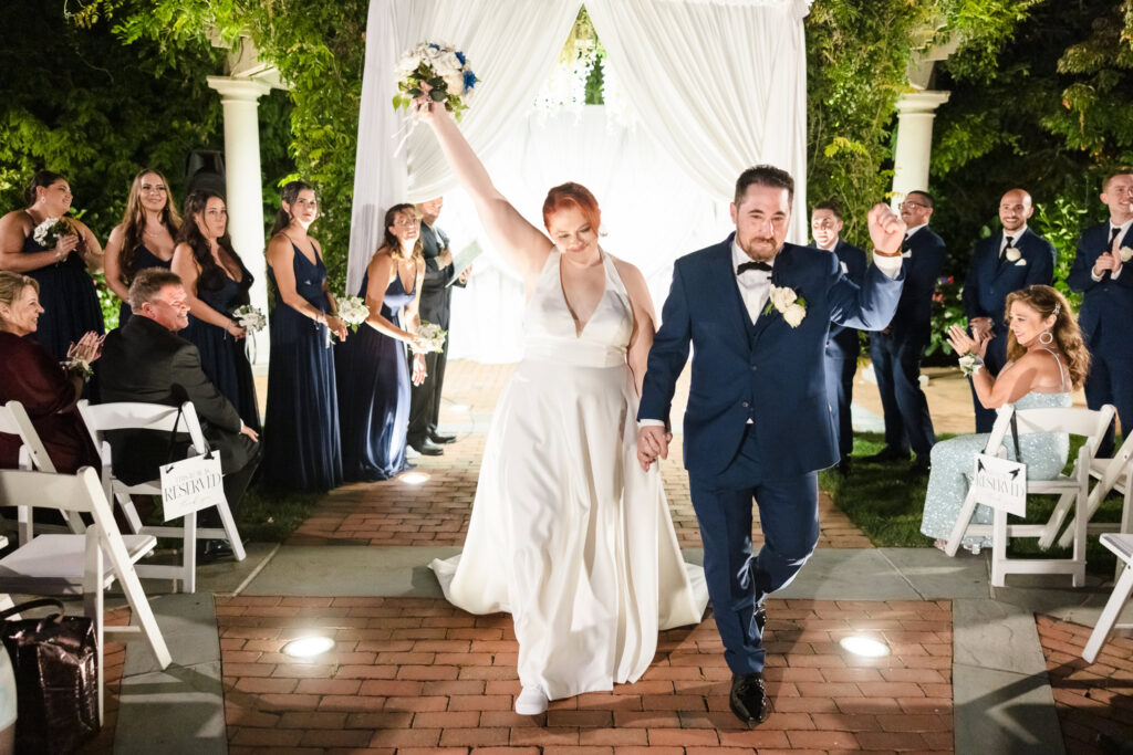 Just married couple walks joyfully down the aisle at their Crest Hollow Country Club wedding, surrounded by cheering guests and elegant garden columns.