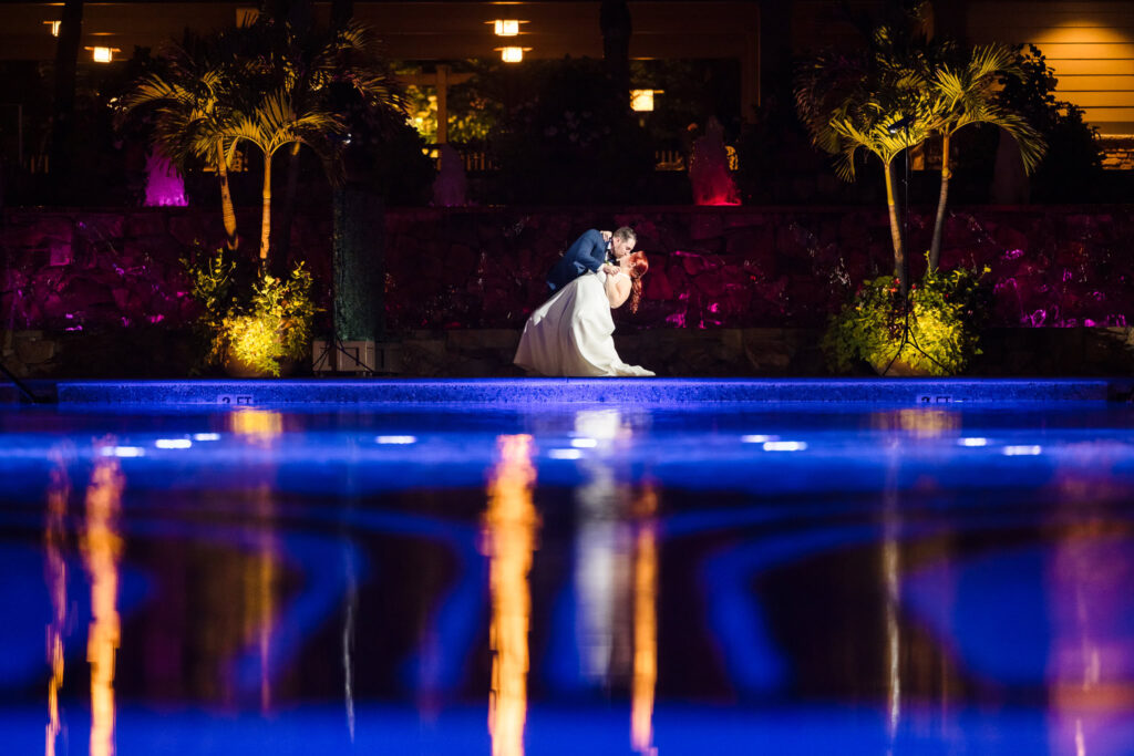 Bride and groom share a romantic kiss beside the illuminated pool at Crest Hollow Country Club, with vibrant reflections and tropical palms framing the night scene.

