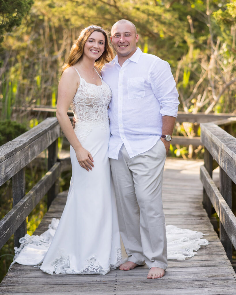 Bride and groom standing barefoot together on a rustic boardwalk surrounded by woods, smiling at the camera.