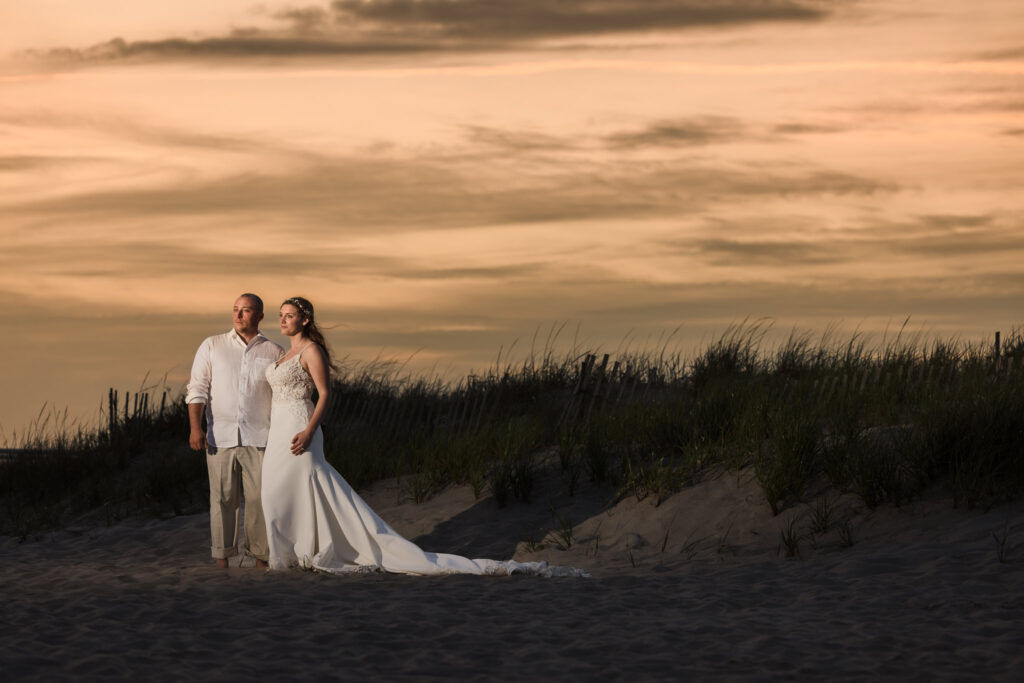 Bride and groom standing barefoot on a beach at sunset, surrounded by dune grass and a golden sky.