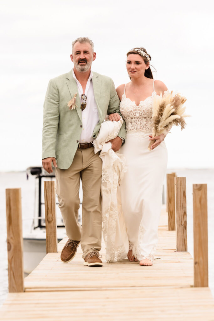 Bride walking down the dock arm-in-arm with her father, holding a bouquet of dried florals.