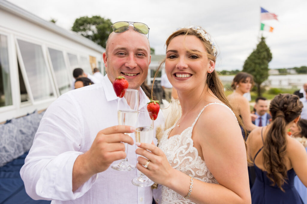 Bride and groom smiling at the camera while toasting with champagne glasses garnished with strawberries.