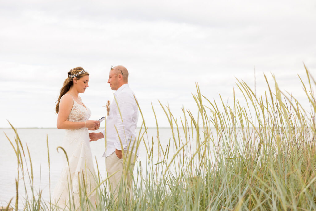 Bride and groom standing in tall grass by the water, exchanging vows during an intimate beachside ceremony.
