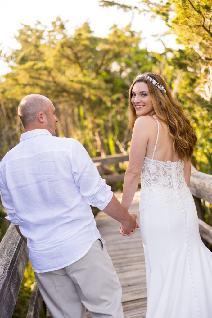 Bride looking back smiling while holding her groom’s hand on a tree-lined boardwalk.