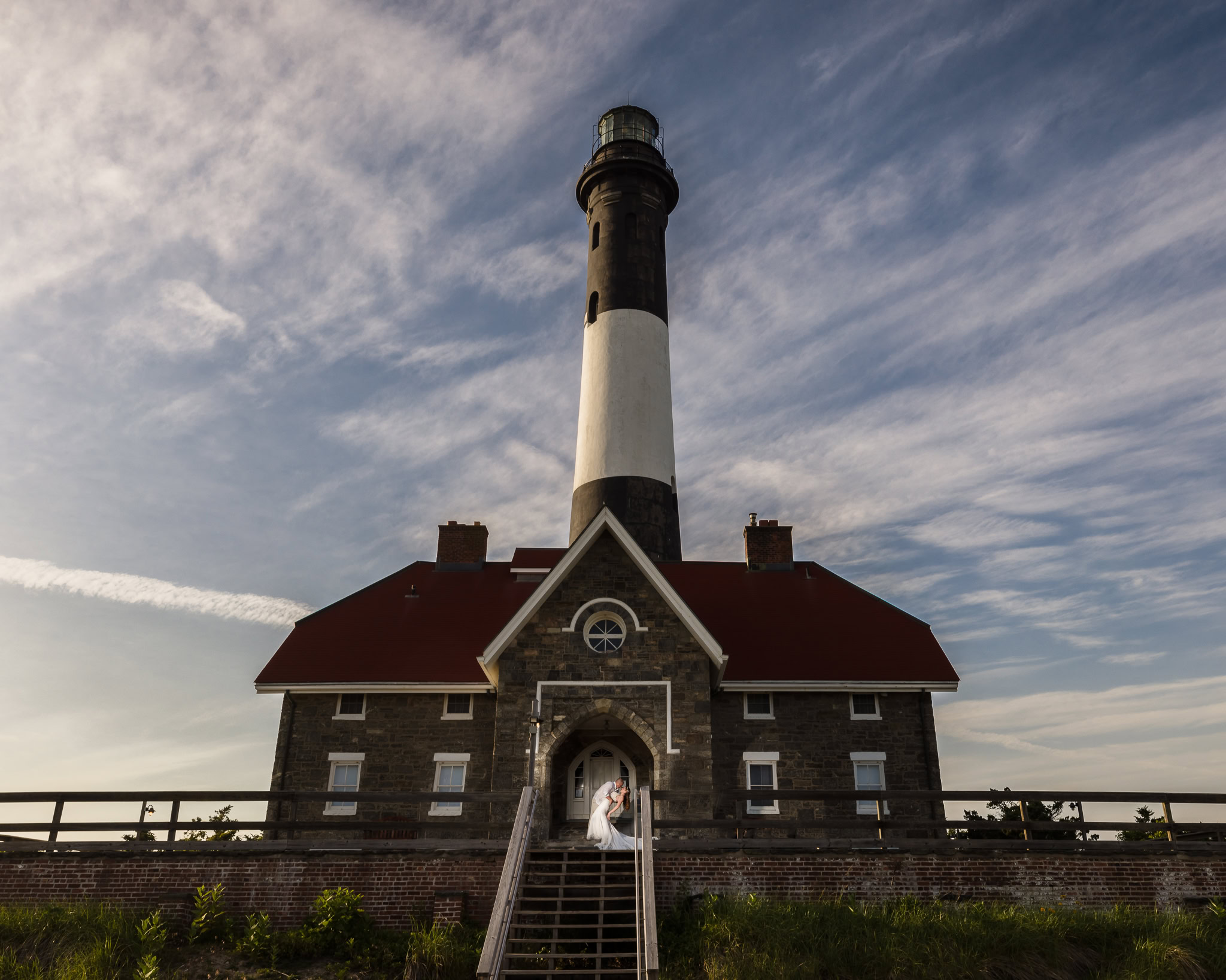 Bride and groom standing on the steps of a historic lighthouse beneath a dramatic sky.