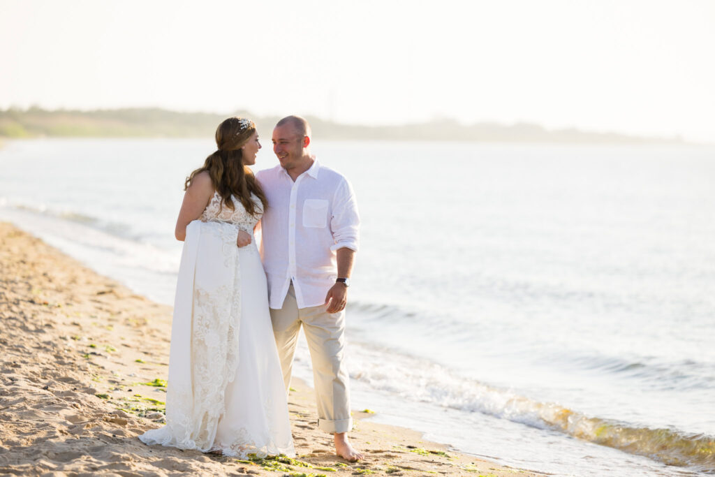 Bride and groom walking hand-in-hand along the shoreline, softly lit by afternoon sun.