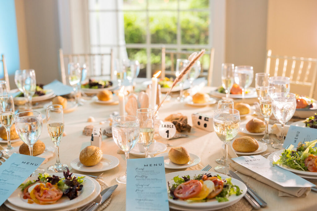 Elegant wedding reception table setting at East Wind Long Island, featuring fresh salads, champagne flutes, and glowing candlelight in a sunlit room.

