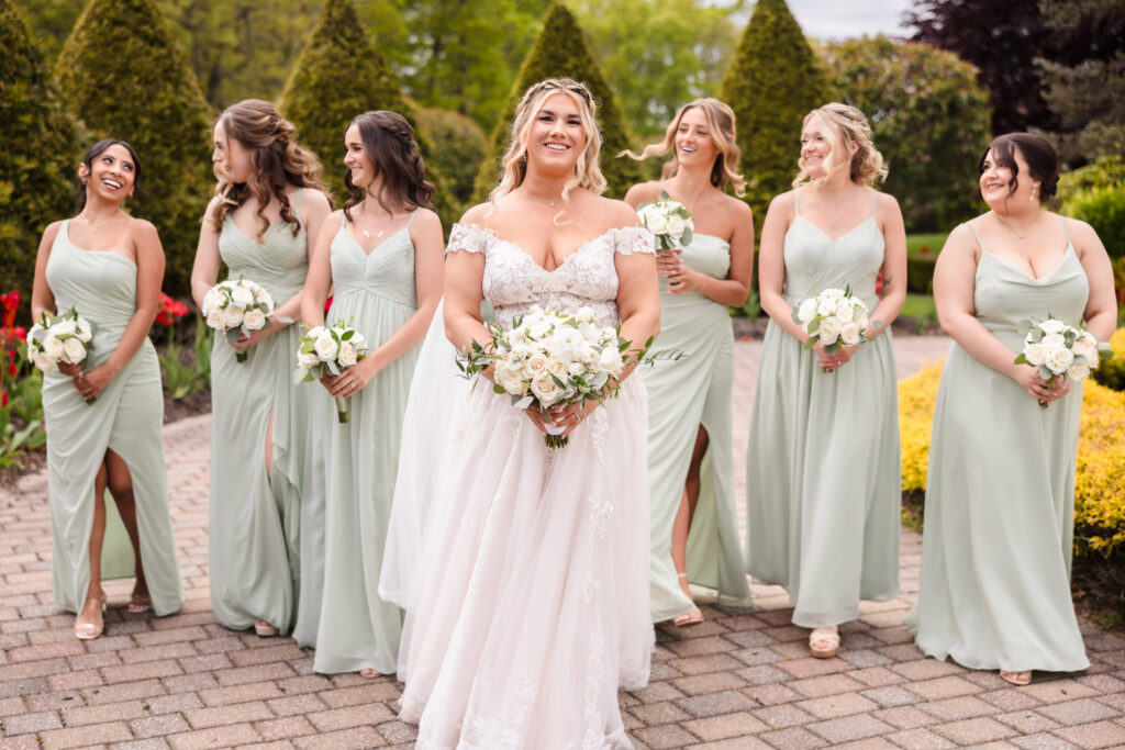Bride walks joyfully with her bridesmaids in sage green dresses through the manicured garden paths at East Wind Long Island.

