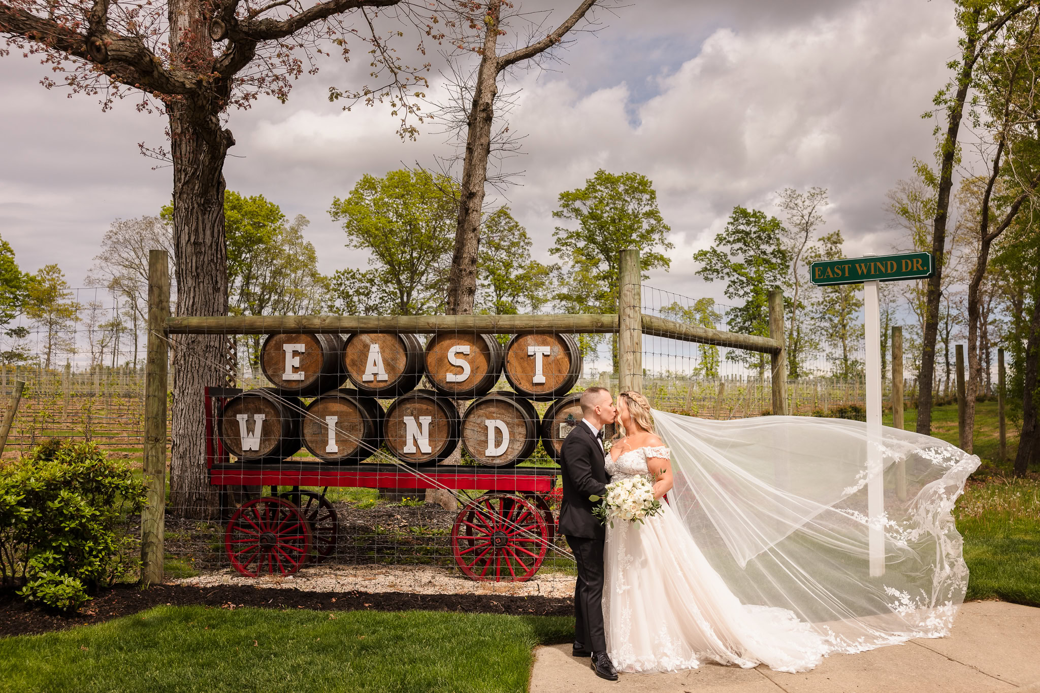 Bride and groom kiss in front of East Wind Long Island’s iconic wine barrel sign, surrounded by spring foliage and a sweeping veil.