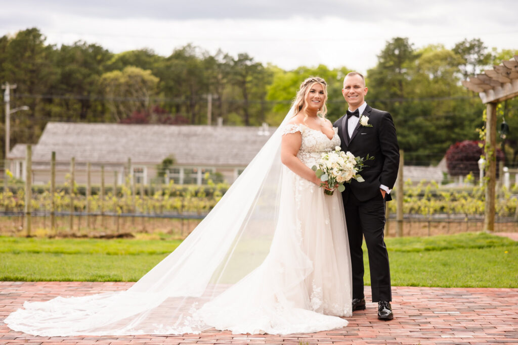 Bride and groom pose elegantly in front of the vineyard at East Wind Long Island, her long veil flowing behind them on a brick path.


