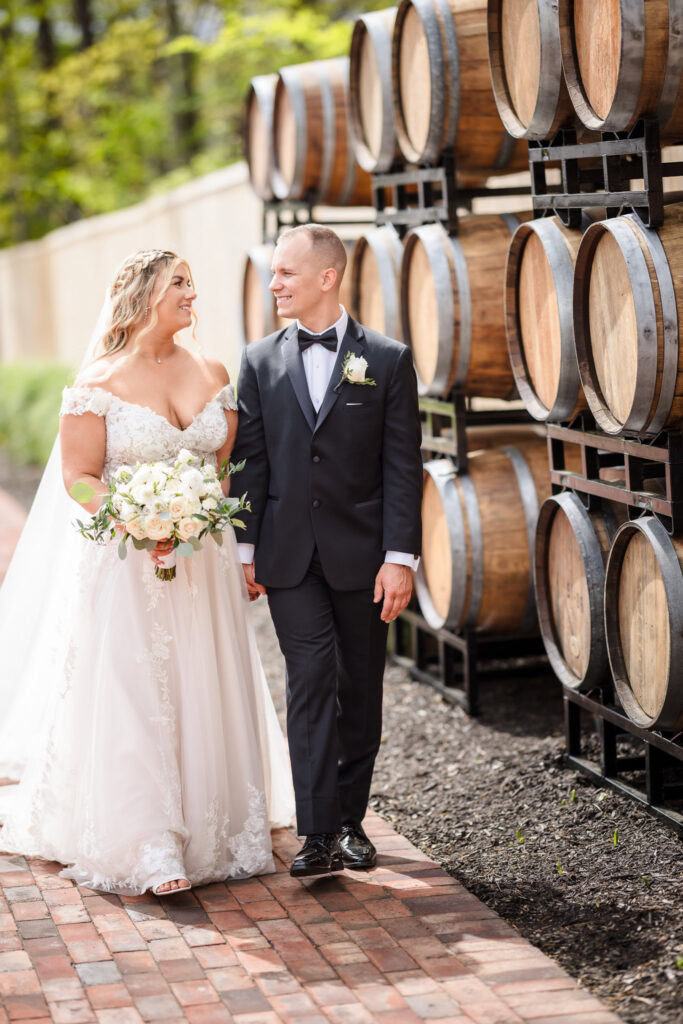 Bride and groom walk hand-in-hand past stacked wine barrels at East Wind Long Island, sharing a quiet moment on the brick path.

