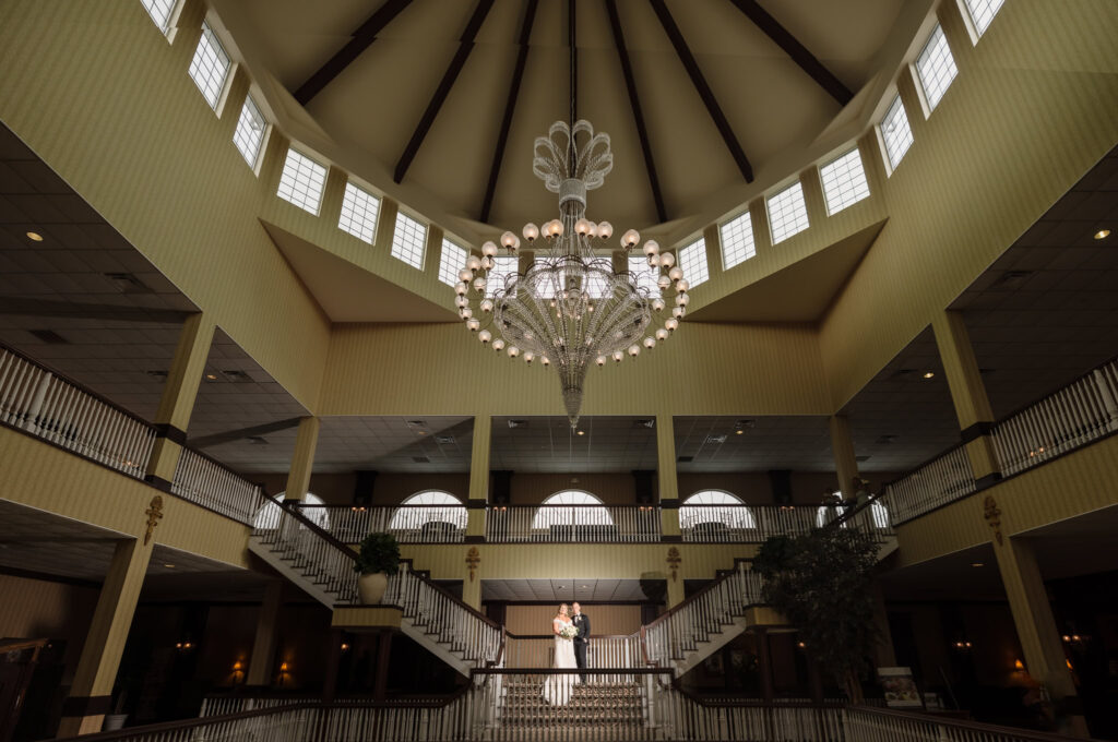 Bride stands elegantly at the top of a grand staircase beneath East Wind Long Island’s iconic chandelier and vaulted ceiling.

