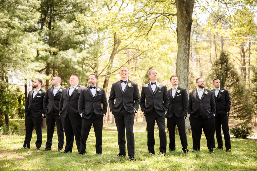 Groom and groomsmen stand confidently in a sunlit grove at East Wind Long Island, dressed in classic black tuxedos.

