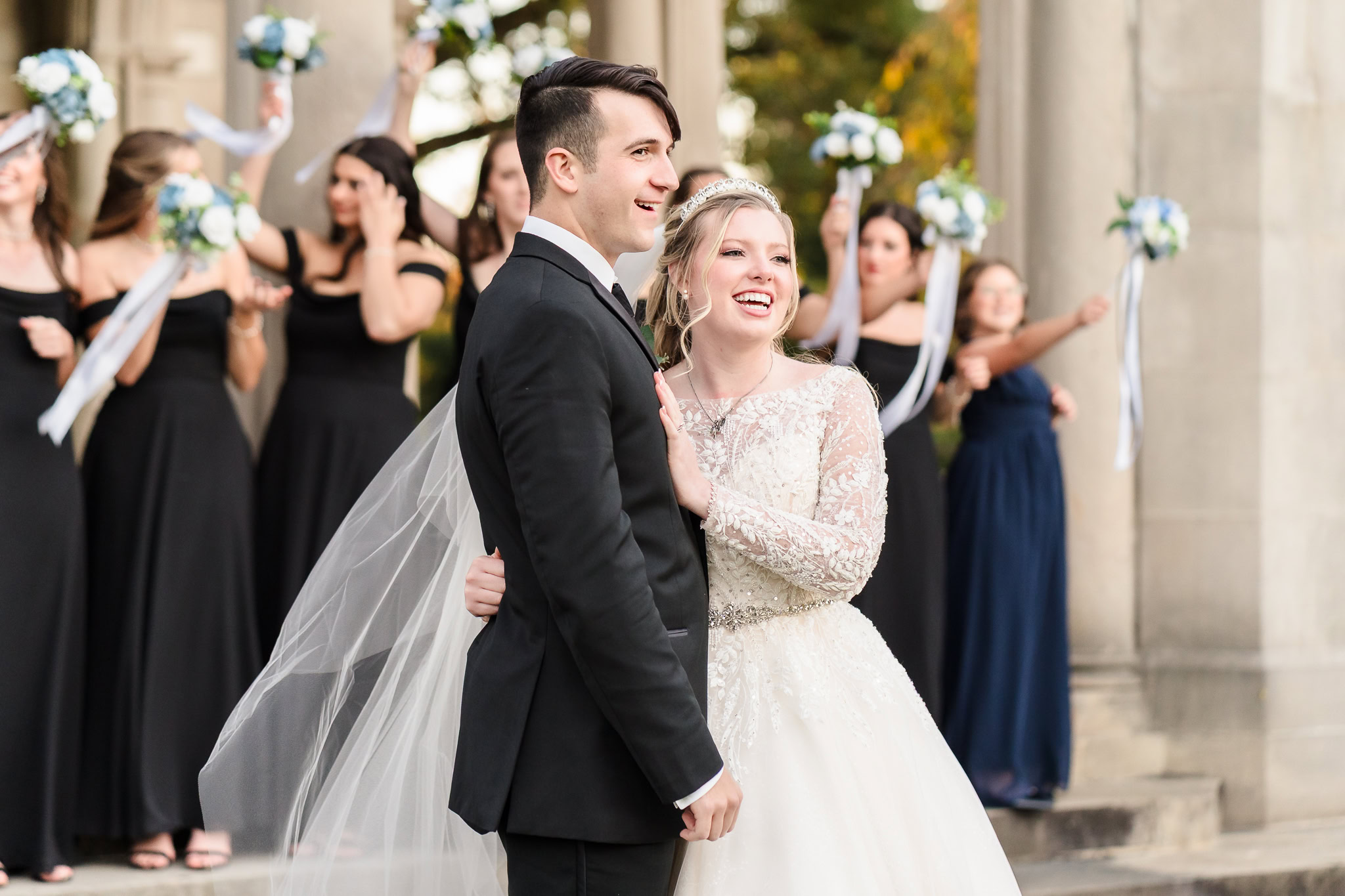 Bride and groom smile joyfully outside Planting Fields Arboretum, with bridesmaids cheering in the background holding white bouquets.