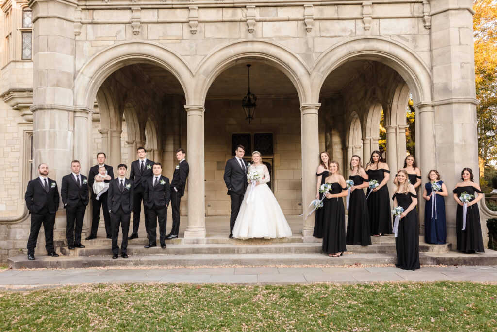 Bride and groom pose with their full wedding party under the stone arches at Planting Fields Arboretum, framed by fall foliage and historic architecture.