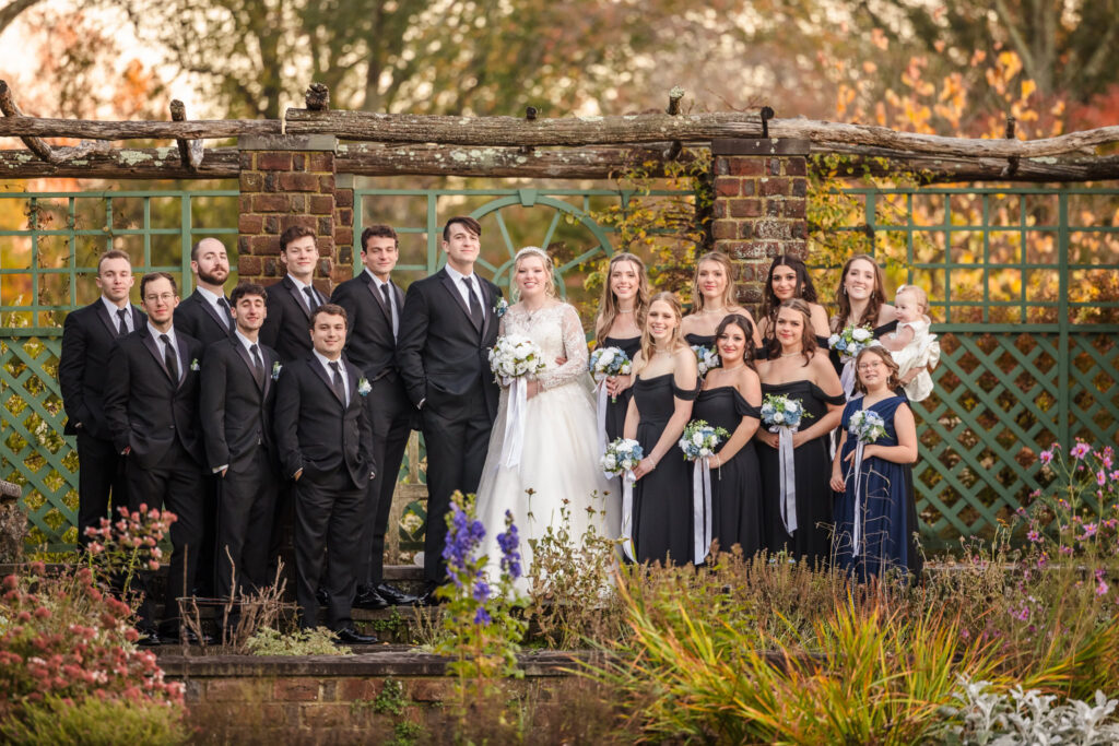 Bride and groom pose with their wedding party in front of a garden trellis surrounded by fall foliage and blooming flowers at Planting Fields.