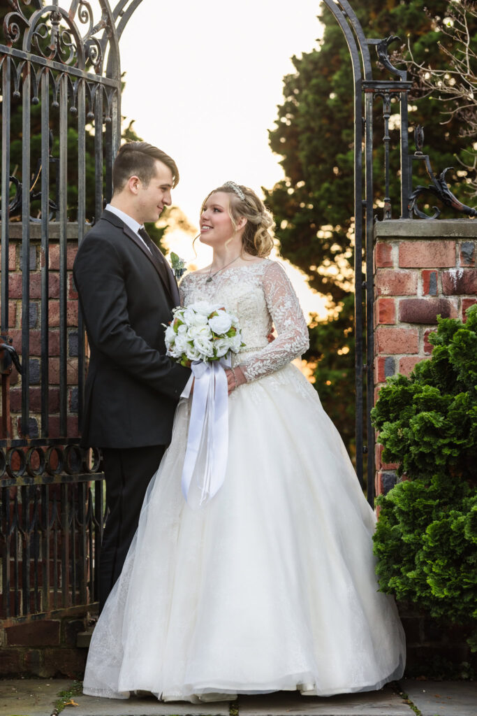 Bride and groom stand in front of an open wrought iron gate framed by brick and greenery, sharing a quiet moment.