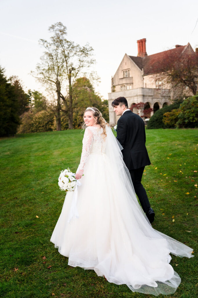 Bride and groom walk hand-in-hand on the lawn with a historic mansion in the background at Planting Fields Arboretum.