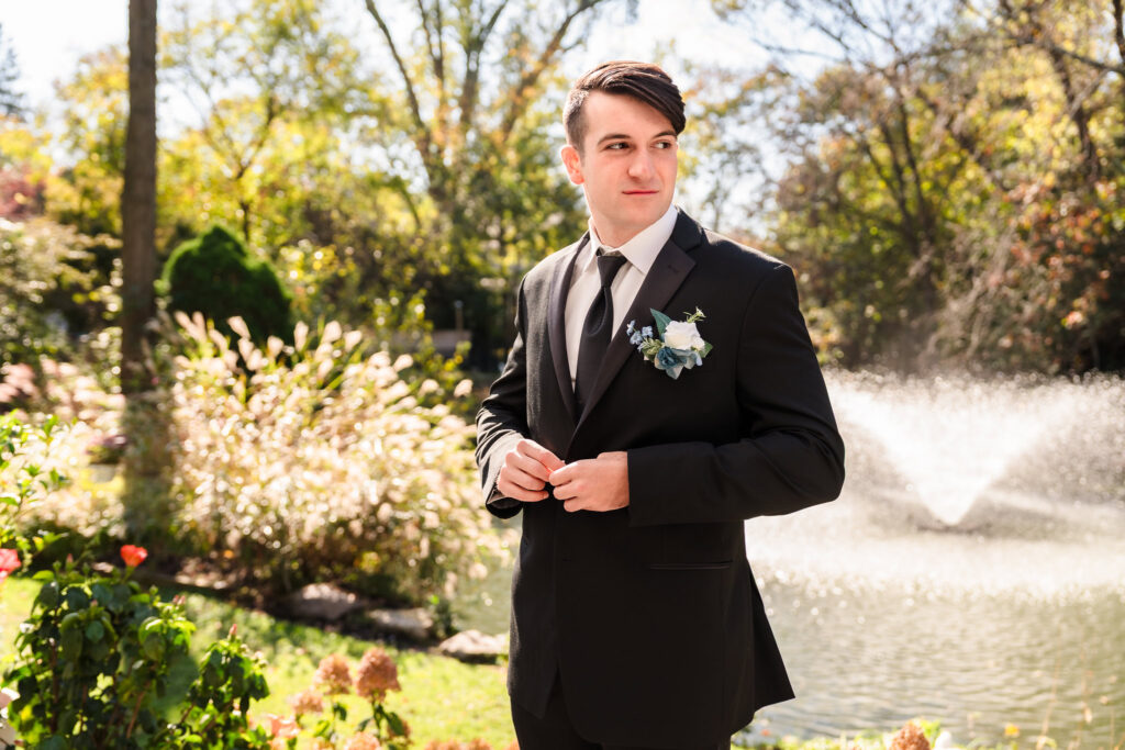 Groom adjusts his cuff while standing outside in sunlight, wearing a black suit and white boutonniere.