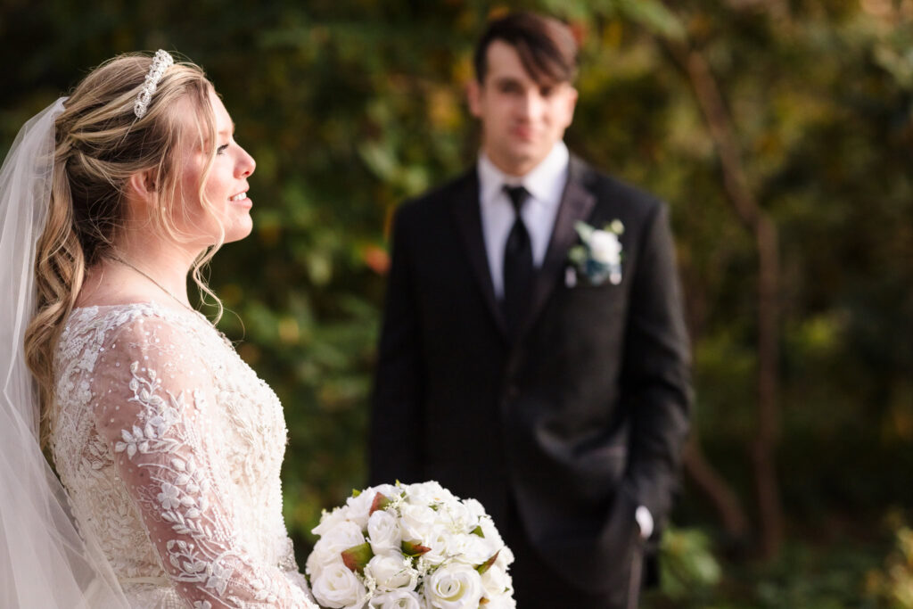 Bride holds a white bouquet and smiles softly in the foreground, with the groom blurred in the background among autumn trees at Planting Fields Arboretum.